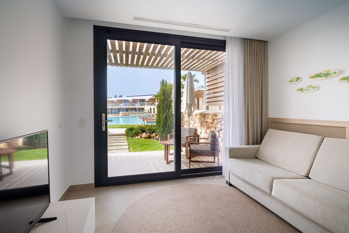 Una habitación de hotel moderna con un amplio ventanal corredero que da a una zona de piscina, un sofá y un televisor de pantalla plana.