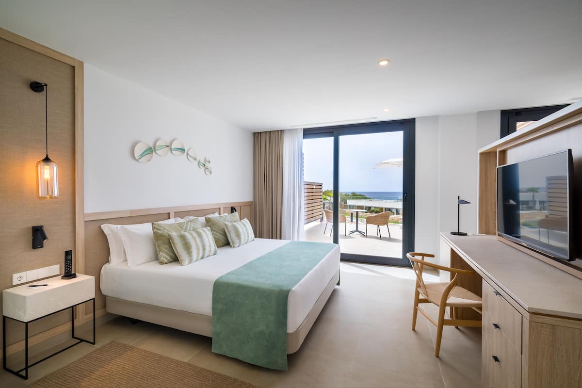 Una habitación de hotel moderna con una cama grande, un escritorio y un televisor. Cuenta con un balcón con vistas al océano.