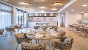 Un amplio comedor con mesas redondas dispuestas para una comida, con un diseño de techo moderno y luces empotradas, y una cocina visible al fondo.
