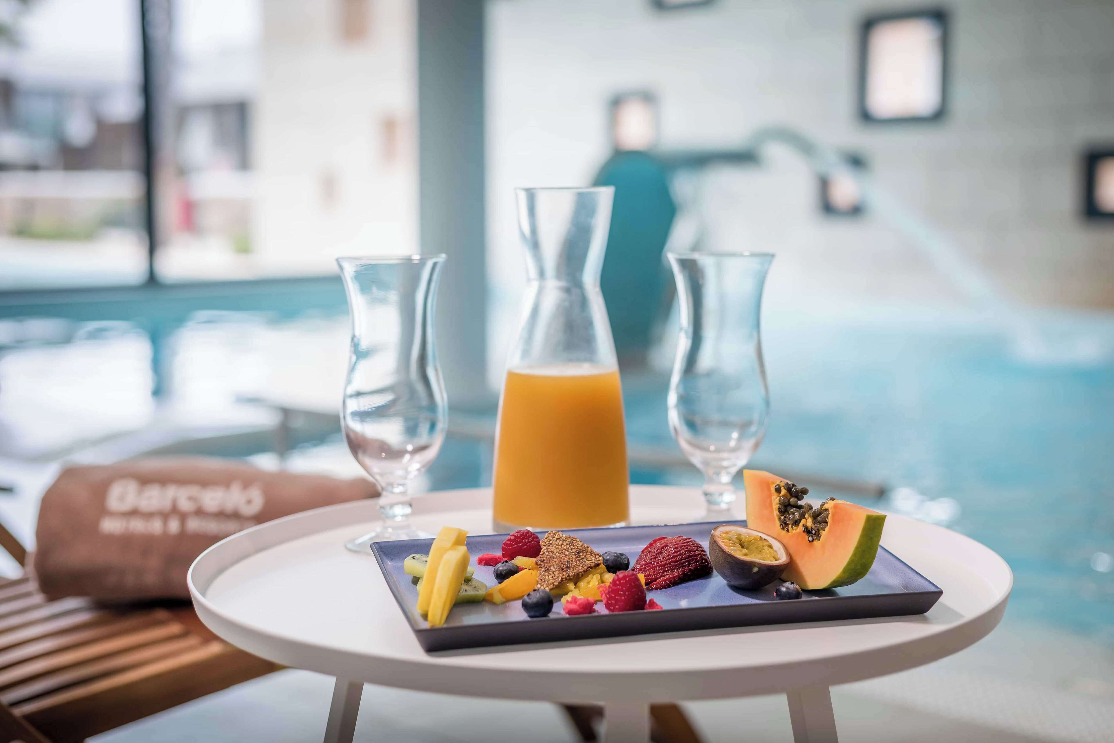 A breakfast setting with a fruit platter, two glasses of orange juice, and a bottle of juice on a table by a pool.