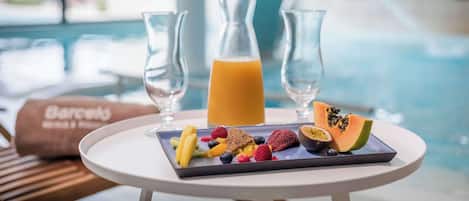 Un desayuno con una bandeja de frutas, dos vasos de zumo de naranja y una botella de zumo en una mesa junto a una piscina.