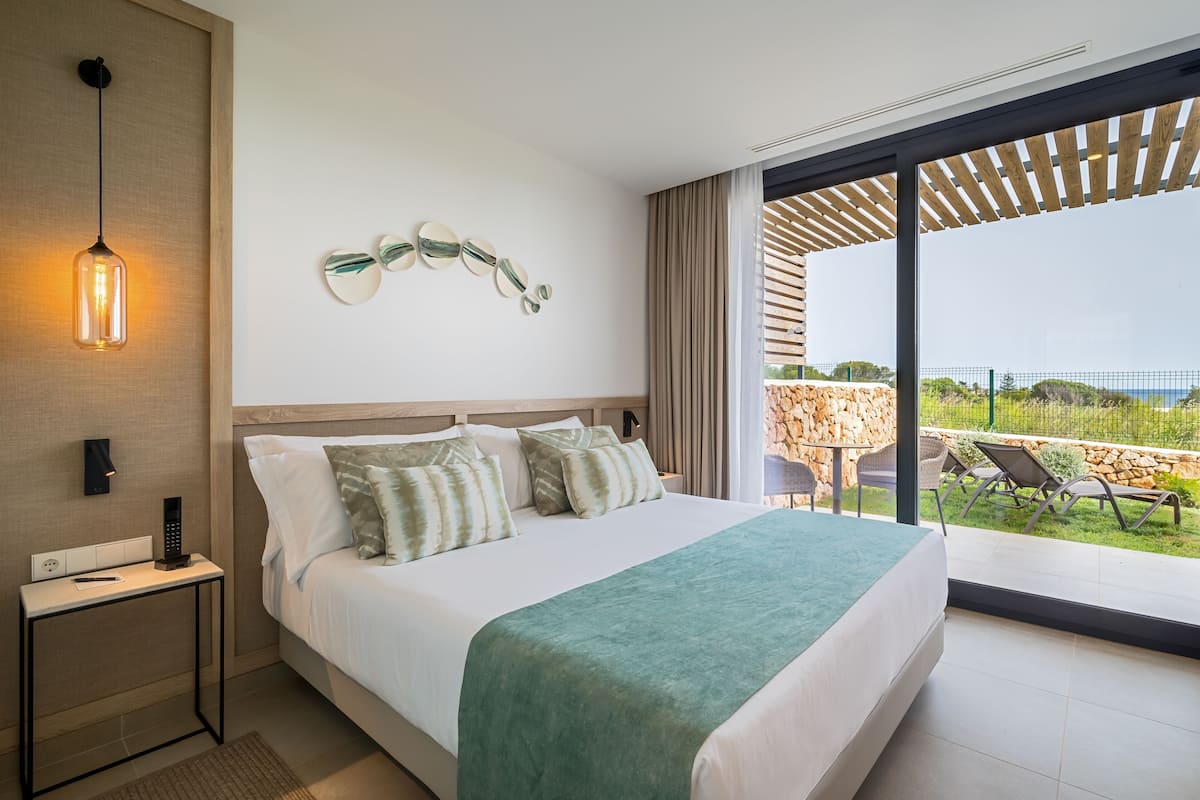 Una habitación de hotel moderna con una cama grande, una mesita de noche con un control remoto, una lámpara de pared y vistas al patio exterior.