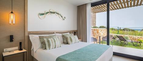 A modern hotel room with a large bed, a bedside table with a remote control, a wall-mounted light fixture, and a view of the outdoor patio with seating.