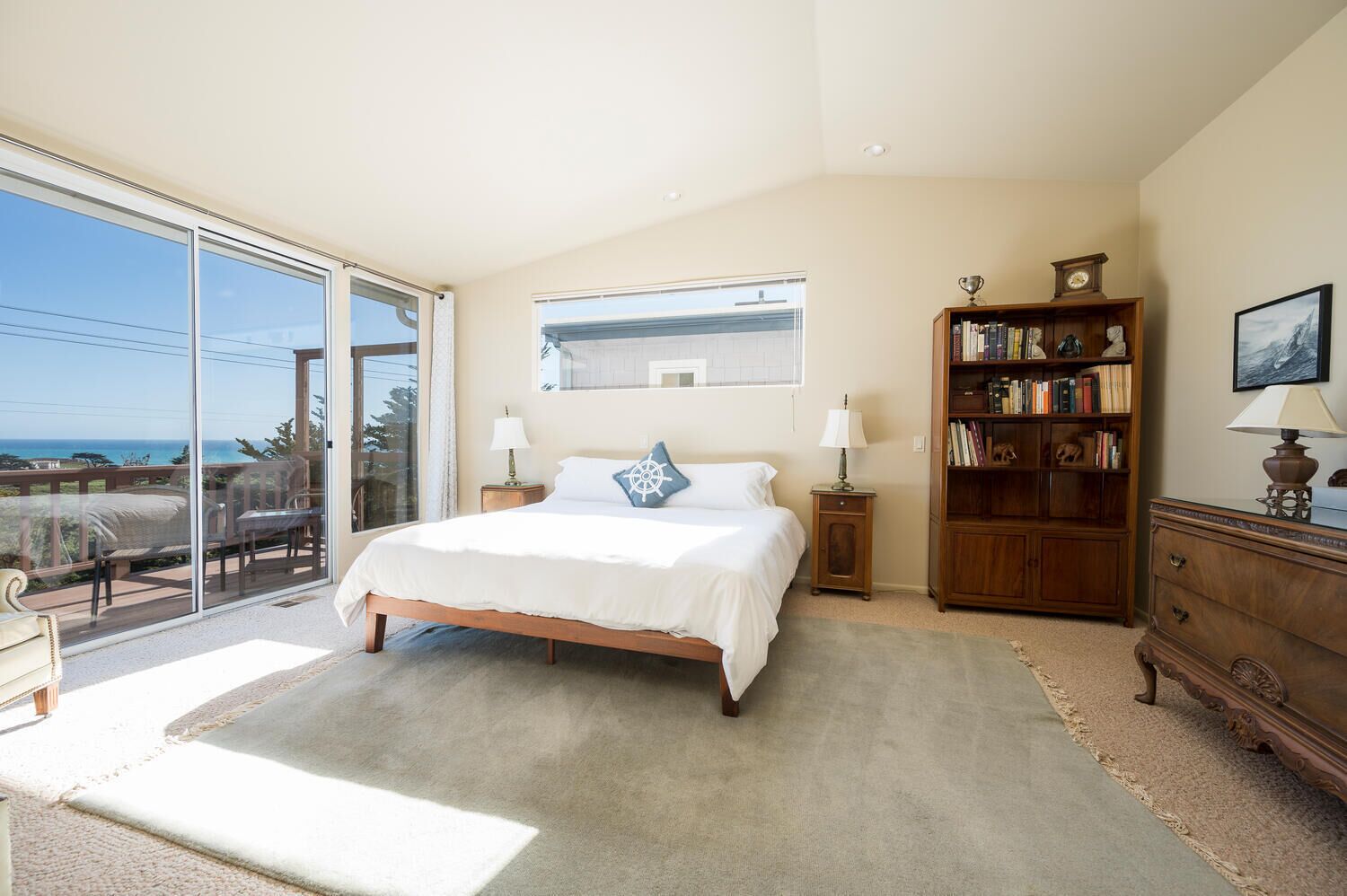 A bedroom with a large bed, a wooden nightstand, a bookshelf, and a view of the ocean through a sliding glass door.