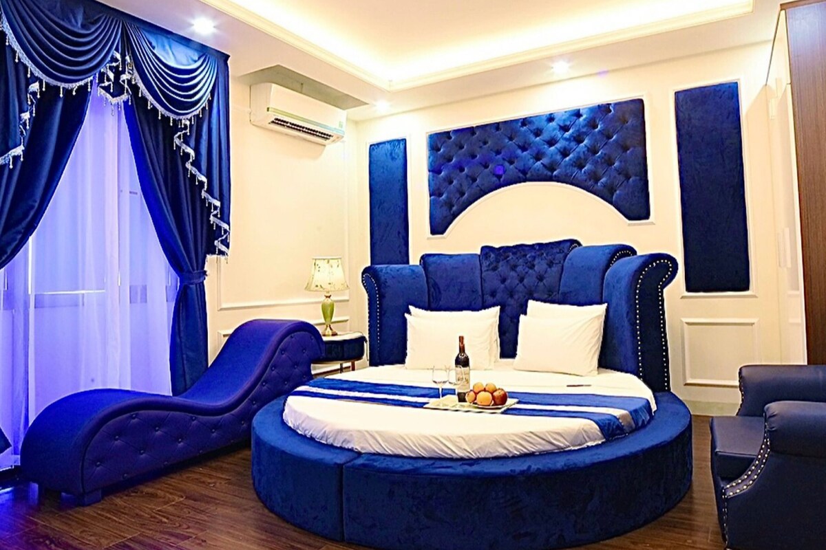 A circular bed with a tufted headboard, a bedside table with a lamp, and a chair.