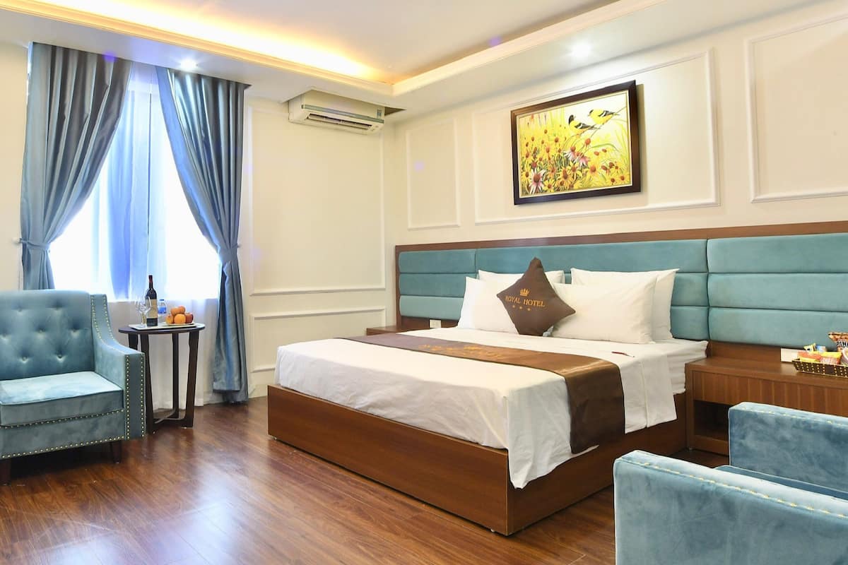 Ein Hotelzimmer mit einem großen Bett, einem blauen Sessel, einem kleinen Tisch mit einer Flasche und Obst sowie einem Gemälde an der Wand.