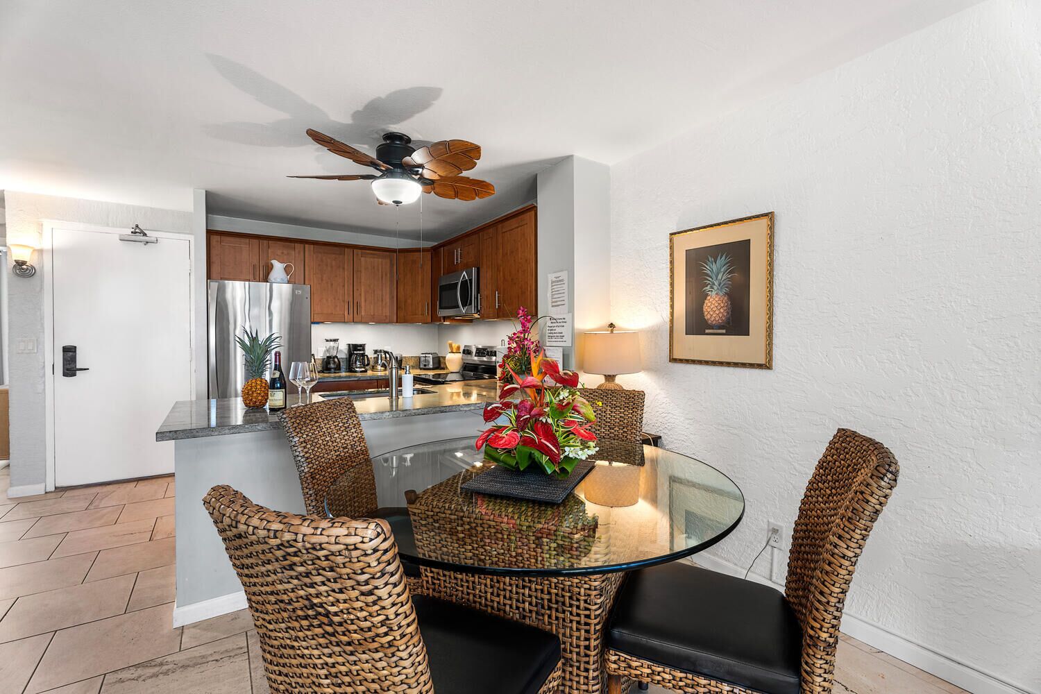 A modern kitchen with wooden cabinets, a stainless steel refrigerator, and a dining area with wicker chairs and a glass table.