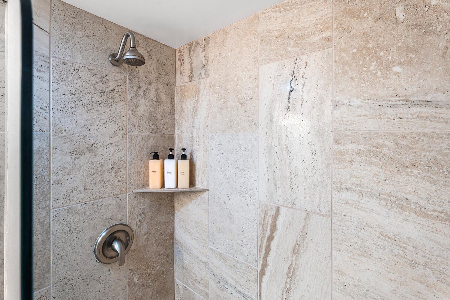 Un espace douche équipé d’une pomme de douche à effet pluie, d’une étagère avec trois bouteilles et de toilettes à chasse d’eau.