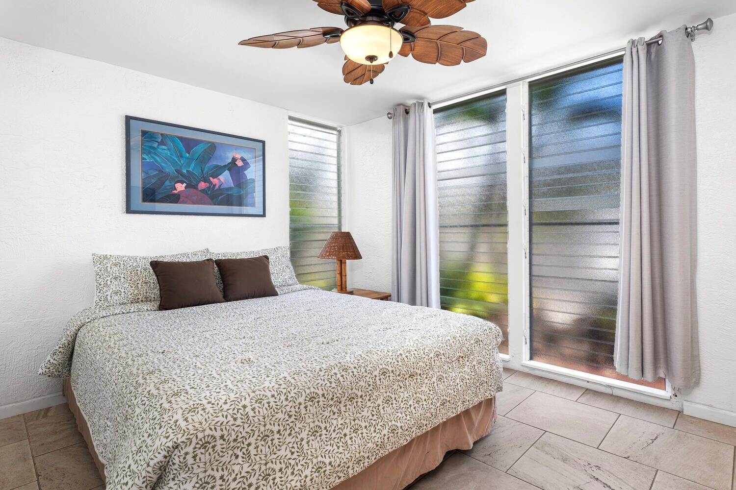 Un dormitorio con cama, ventilador de techo, ventana con cortinas y un cuadro en la pared.