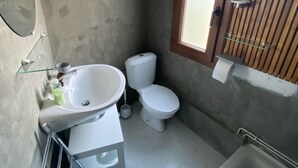 Un baño con lavamanos, inodoro y bañera blancos. Hay una ventana con persianas de madera y un perchero.