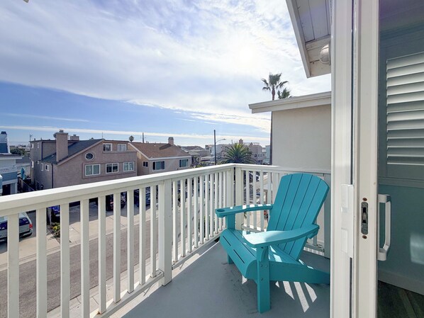 A balcony with a white railing, two turquoise Adirondack chairs, and a view of a residential area with houses and a palm tree. - 139H - Absolute Beach (Oxnard)