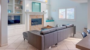 A living room with a fireplace, a television, a sofa, and a window with blinds. - 139H - Absolute Beach (Oxnard)