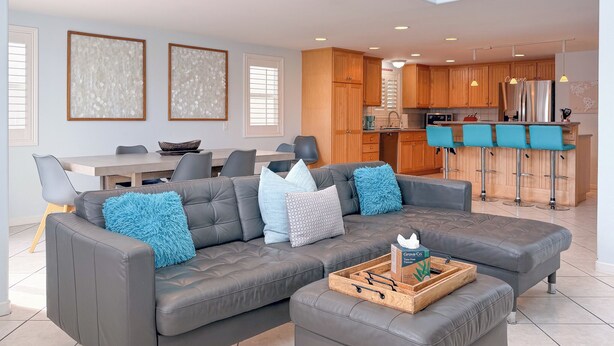 A modern living room with a grey sectional sofa, a wooden coffee table, and a kitchen area with wooden cabinets and a bar counter. - 139H - Absolute Beach (Oxnard)