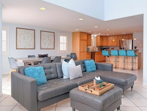 A modern living room with a grey sectional sofa, a wooden coffee table, and a kitchen area with wooden cabinets and a bar counter. - 139H - Absolute Beach (Oxnard)