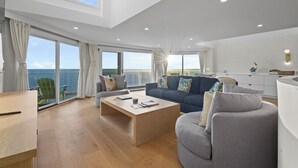 A spacious living room with a large window overlooking the sea, a wooden coffee table, a grey sofa, and a modern kitchen area.