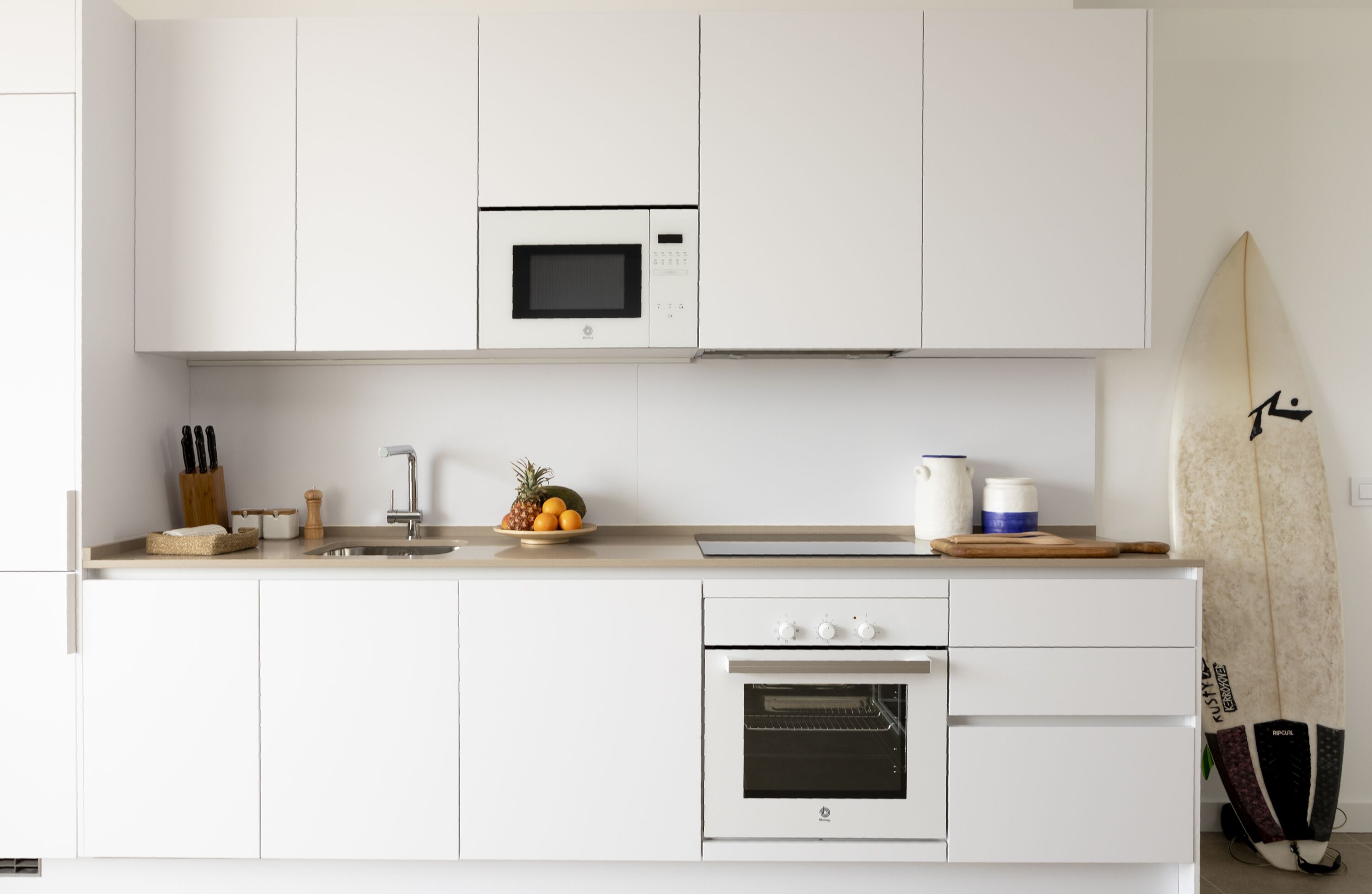 A modern kitchen with white cabinets, a microwave, a sink, and a surfboard leaning against the wall.