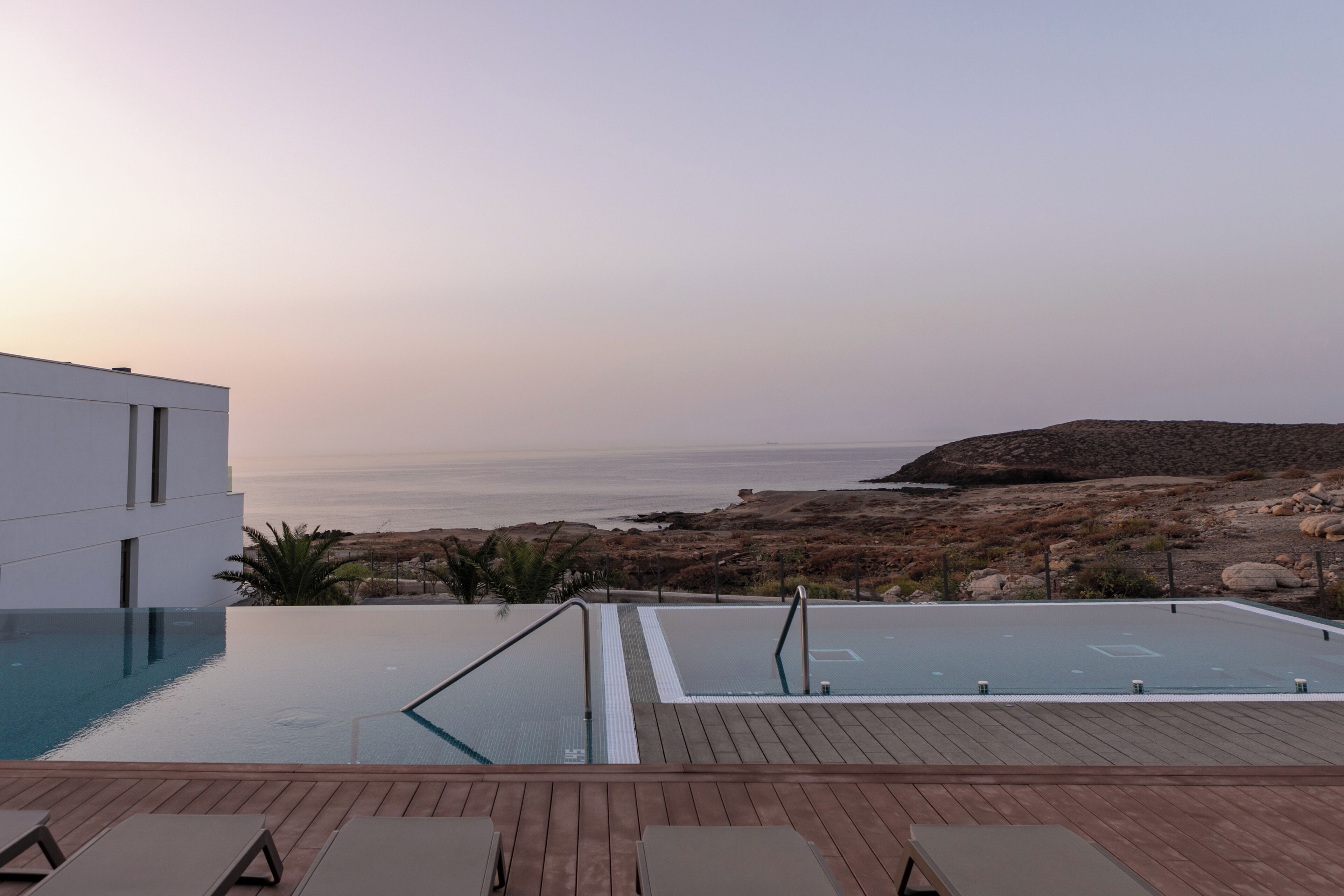 A rooftop infinity pool with a wooden deck and lounge chairs, overlooking a rocky coastline and the sea.
