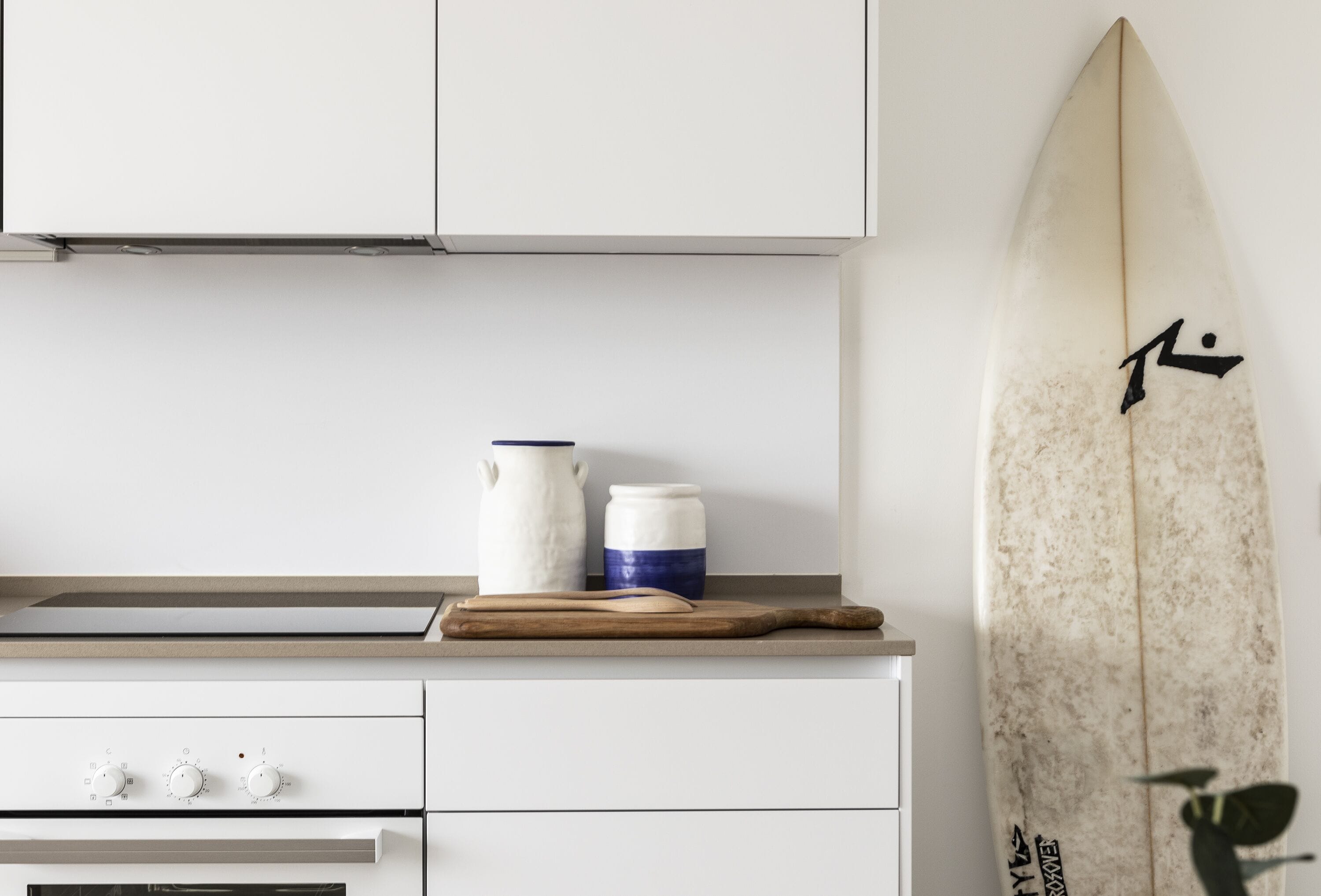 A kitchen with white cabinets, a countertop with a cutting board, a white pitcher, and a blue container. A surfboard with a black symbol is leaning against the wall.