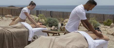 Un espace spa comprenant deux tables de massage, une petite table en bois avec des bouteilles, et une vue sur l’océan.