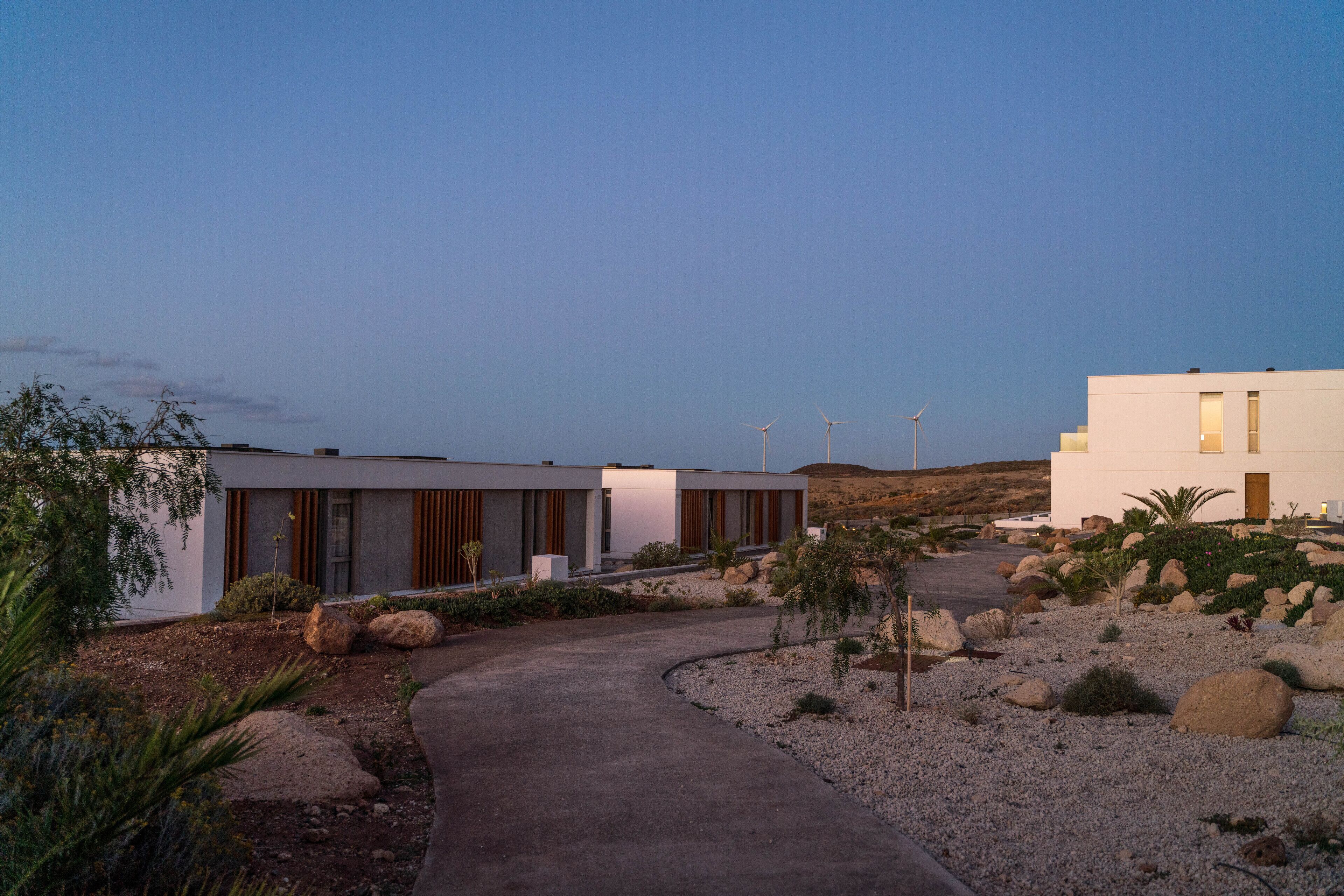 Un bâtiment moderne, avec un chemin menant à une structure blanche, entourée de rochers et d’une végétation clairsemée.