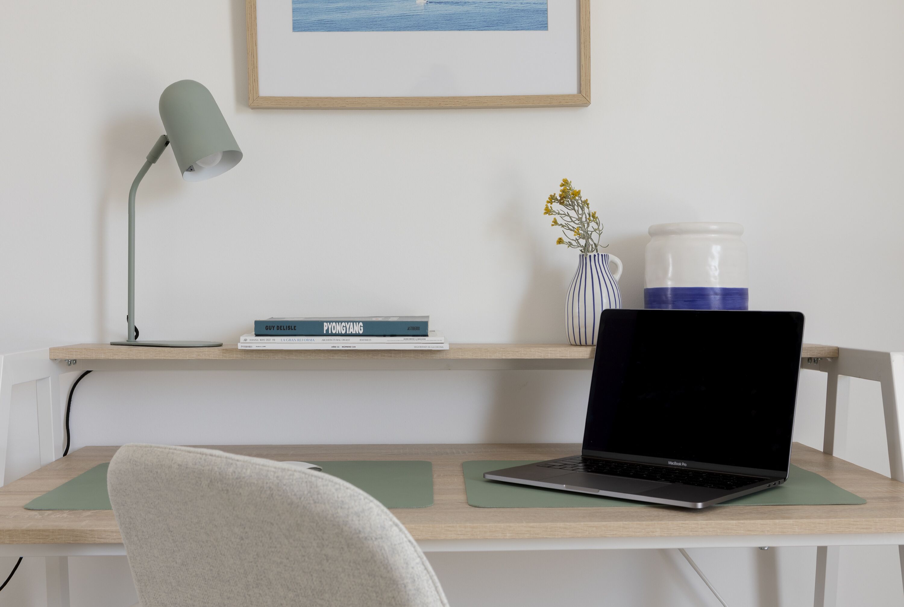 Ein Arbeitsplatz mit Laptop, Lampe, Blumen in einer Vase und einem gerahmten Bild an der Wand.