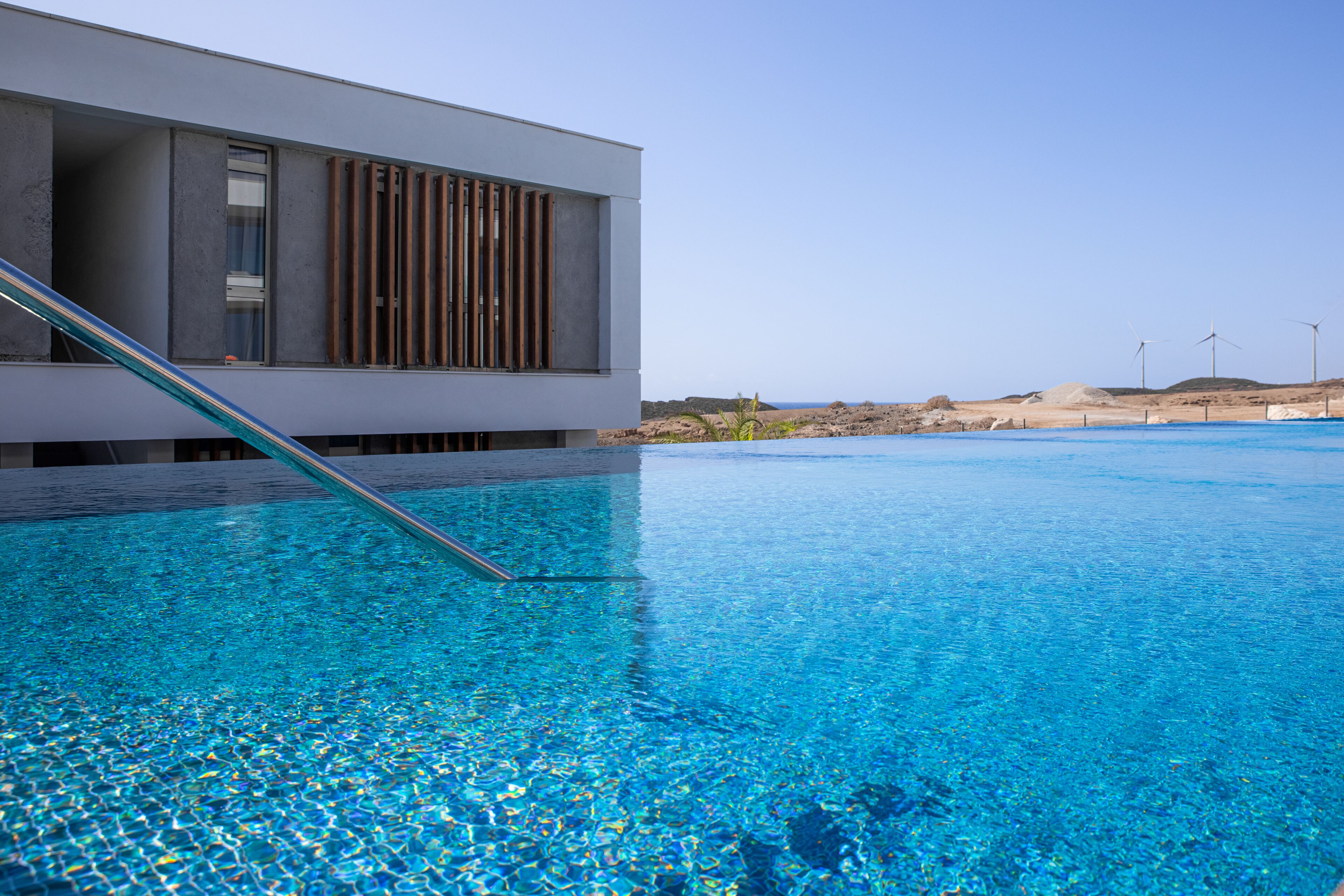 A modern building with a pool, clear blue water, and a view of the beach and wind turbines in the background.