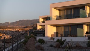 A modern two-story house with a balcony, surrounded by a desert landscape and wind turbines in the background. - Kora Nivaria Beach (Arico)