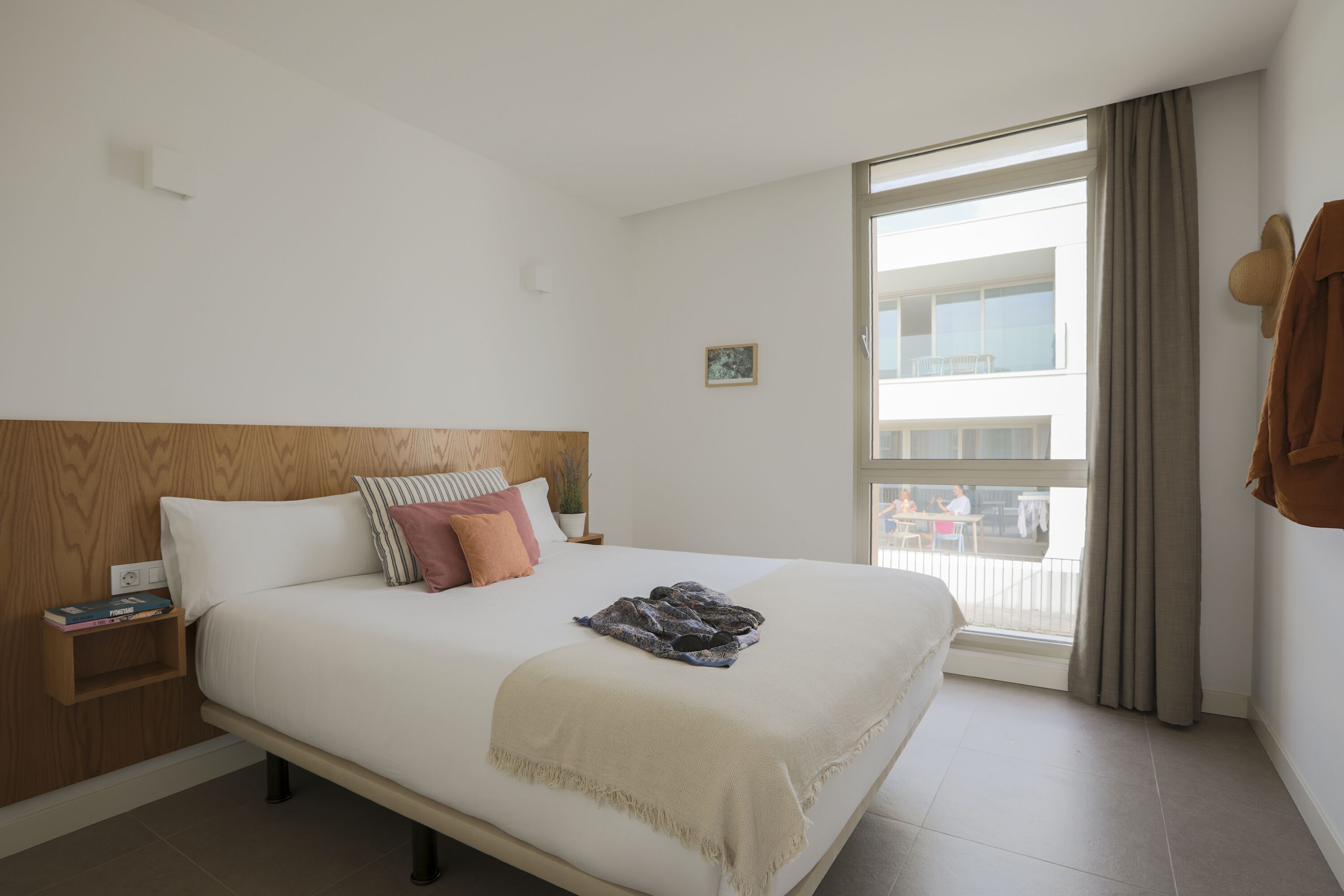 A hotel room with a bed, a nightstand, a window with curtains, and a view of the outside balcony.