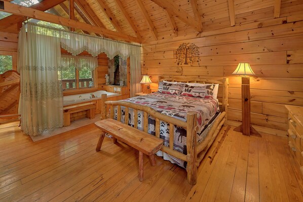 A wooden cabin bedroom with a large bed, a bench, a wooden desk, and a window with curtains.