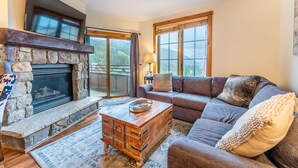 A living room with a stone fireplace, a large flat-screen TV mounted above it, a gray sofa with decorative pillows, and a wooden coffee table. - Overlooks Keystone Base Area, Short Walk to Lifts; Private Balcony with Views (Keystone)
