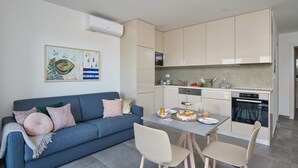 A modern kitchen with a dining area, featuring a blue sofa, a dining table with chairs, and a kitchen island with a sink and stove. - Irena Apartments (Konavle)