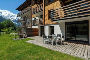 Un bâtiment moderne à plusieurs étages, doté d’une façade en bois, d’une terrasse avec mobilier d’extérieur et offrant une vue sur des montagnes enneigées.