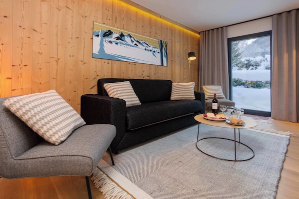 Un salon moderne avec un mur en bois, un canapé noir, des fauteuils gris, un tapis à motifs et une table à manger avec une bouteille et des verres.