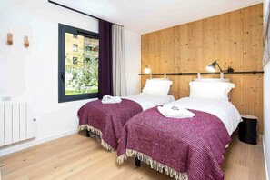 Habitación de hotel con dos camas, ventana con vista a edificios, pared de madera y una lámpara de noche.