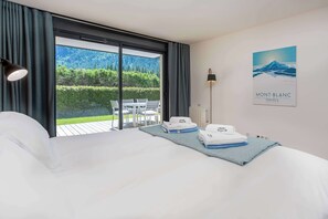 A hotel room with a bed, a view of a garden, a table with chairs, and a painting on the wall.