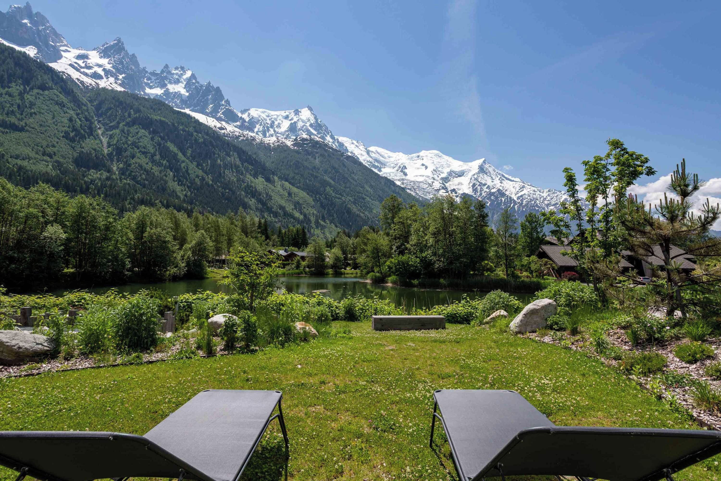 A mountain landscape with a lake, trees, and two lounge chairs facing the view.