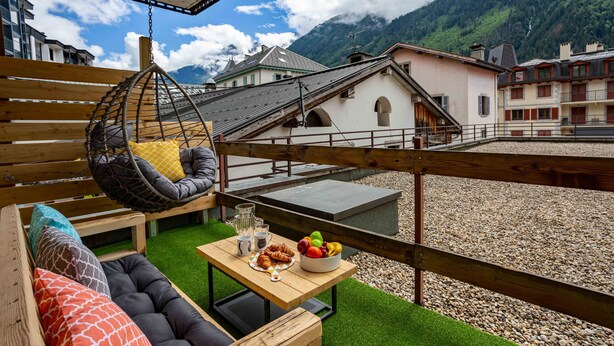 A balcony with a hanging chair, a wooden table with food and drinks, and a view of buildings and mountains.