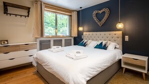 A bedroom with a large bed, a bedside table, a nightstand, a window with curtains, a radiator, and a heart-shaped wall decoration.