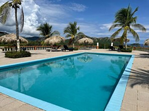 A rectangular swimming pool with a blue edge, surrounded by palm trees and thatched umbrellas, set against a mountainous backdrop. - Cosy 2 bedroom and 2 bath condo walking distance to the beach. (Playas del Coco)