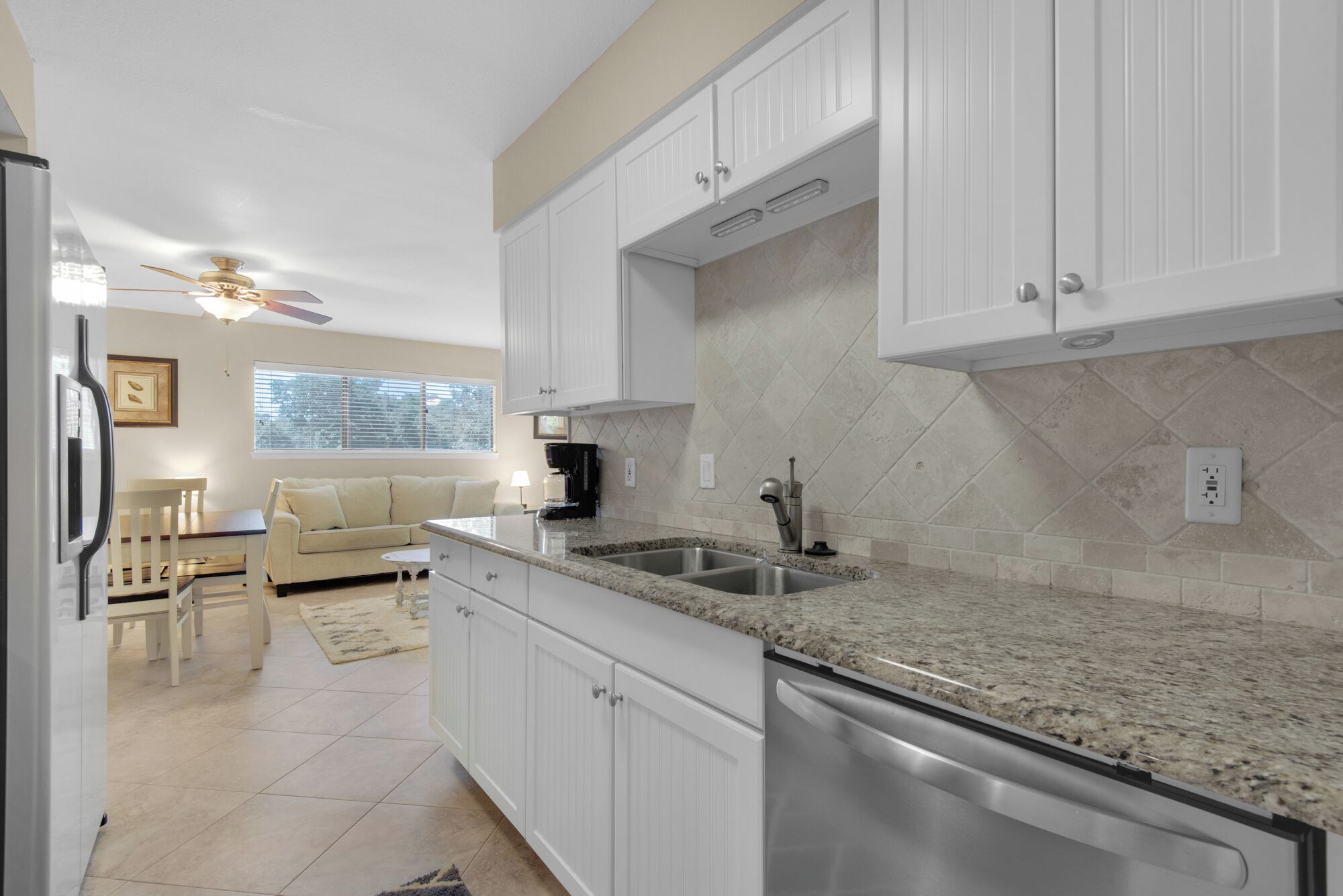 A modern kitchen with white cabinets, granite countertops, and a dishwasher. There is a dining area with a table and chairs, and a ceiling fan above.