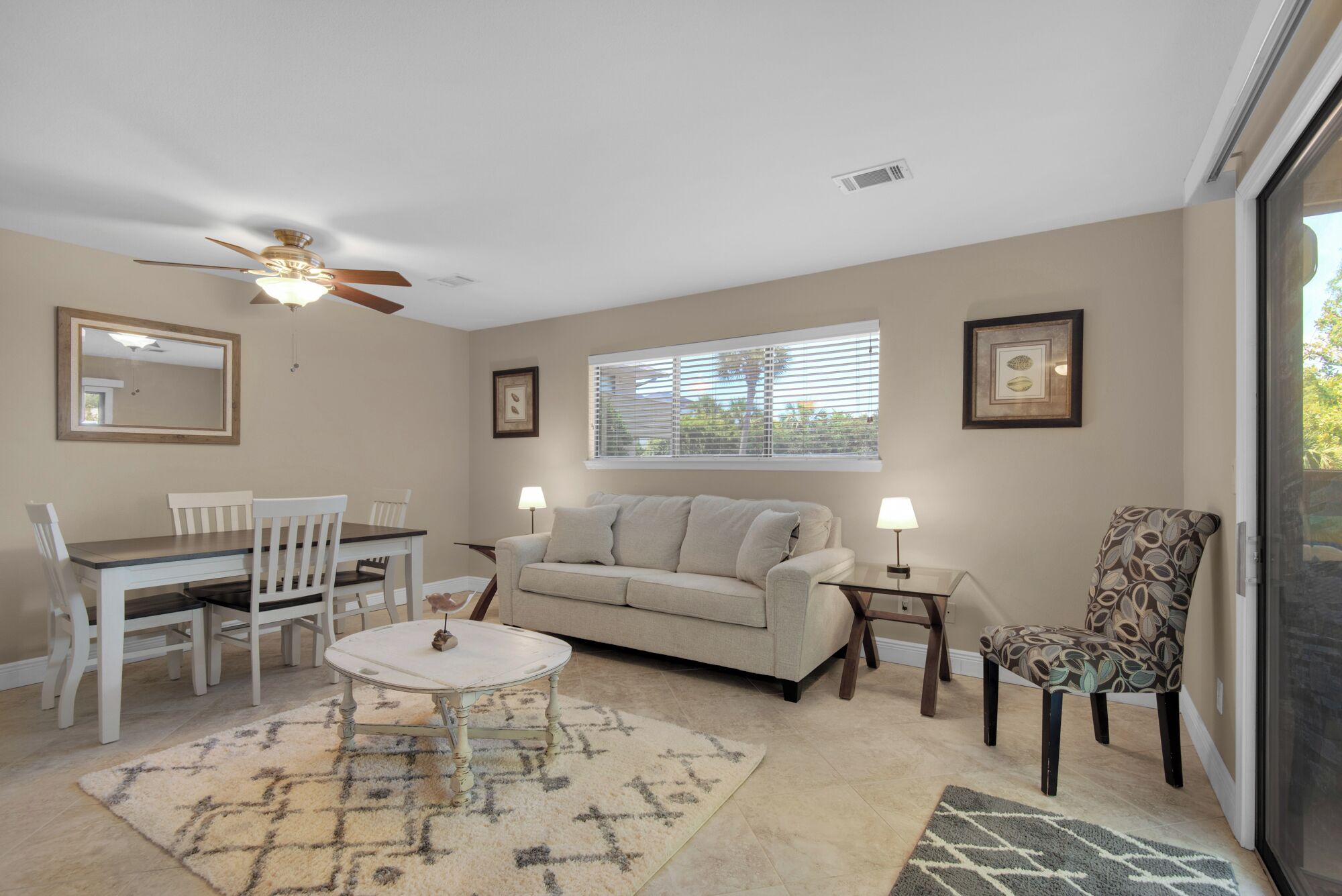 A living room with a dining table, chairs, a sofa, a patterned rug, a ceiling fan, and a window with blinds.