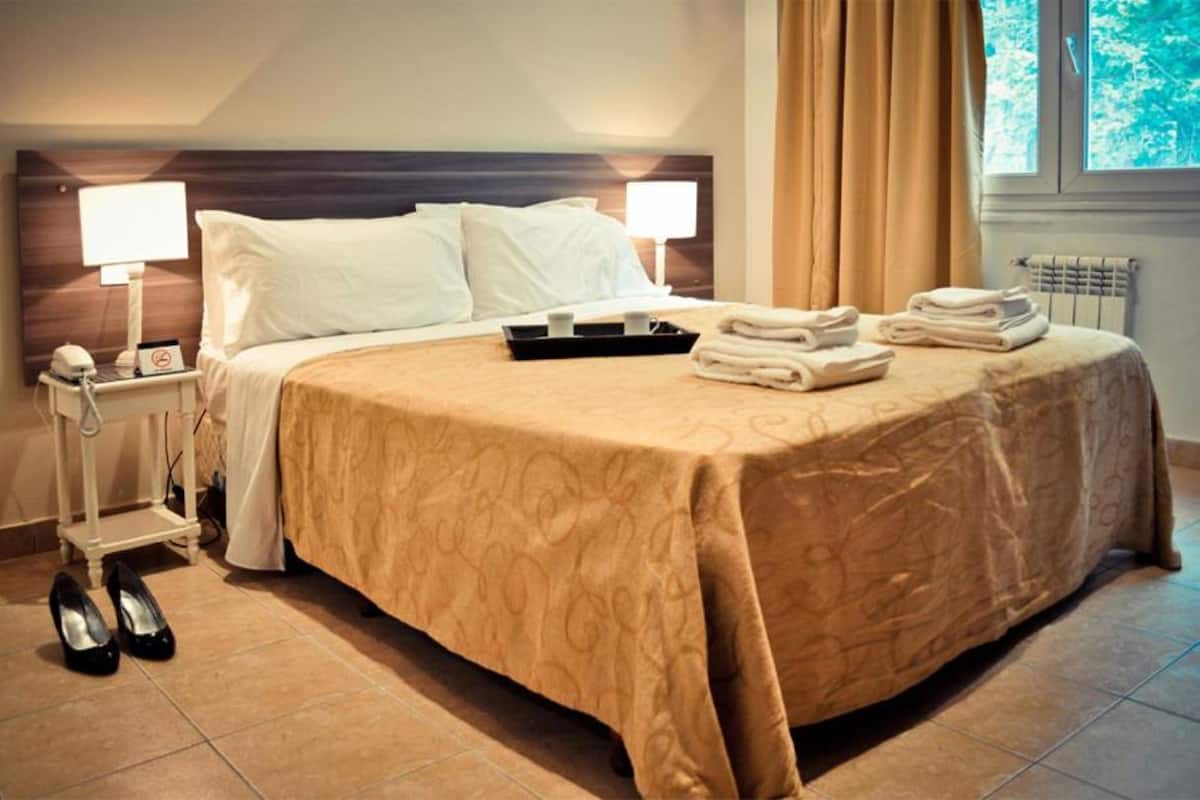 A neatly made bed with a patterned bedspread, two bedside lamps, a tray with towels, and a pair of shoes on the floor.