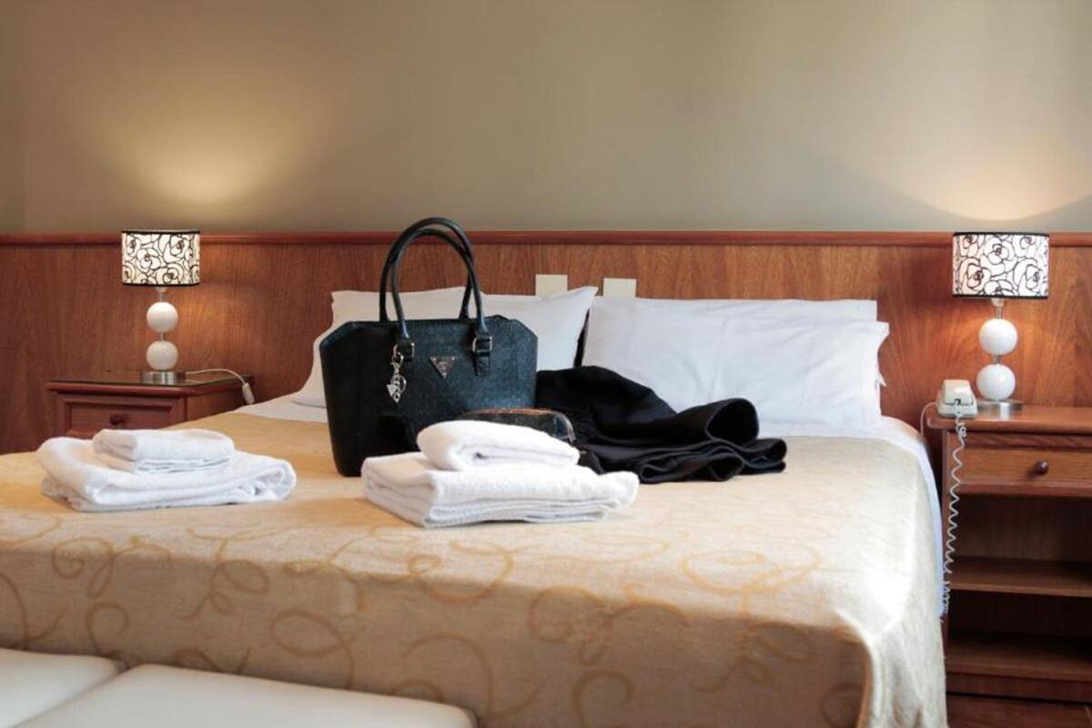 A neatly made bed with a patterned bedspread, a black handbag, folded towels, and a black robe on a wooden headboard.