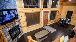 L’intérieur chaleureux d’un chalet avec une cheminée en pierre, un téléviseur, un coin repas comprenant une table ronde et des chaises, et un plafond en bois.