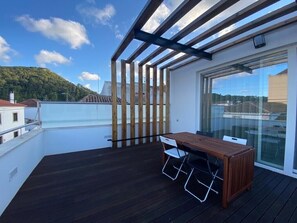 Une terrasse en bois avec une table et des chaises, une porte vitrée donnant sur un balcon, et une vue sur une colline et des bâtiments au loin.