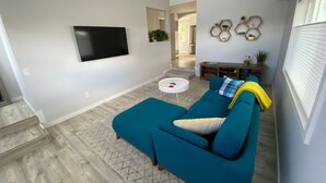 A living room with a teal sofa, a flat-screen TV mounted on the wall, a small white table, and a patterned rug. - Newly Remodeled 4th Street Work-Friendly Home by RedAwning (Bend)