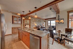 A spacious kitchen with granite countertops, a built-in island, and a stone backsplash. There is a dining area with a glass table and chairs, and a cozy seating area with a sofa and a fireplace. - Cozens Pointe I-101 2 Bedroom Condo by RedAwning (Fraser)