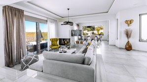 Una sala moderna con un sofá, una mesa de centro y un televisor. Hay una zona de comedor con sillas y una mesa, y un ventanal con cortinas.