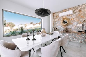 A dining area with a white table, white chairs, a hanging light fixture, a stone wall with a mirror, and a large window with a view of a landscape.