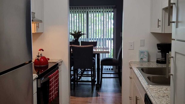 Een kleine keuken met een koelkast, fornuis en wastafel. Er is een eethoek met een tafel en stoelen.